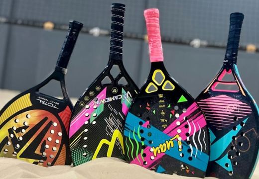 Raquetes e bolas de beach tennis na areia, com a quadra coberta do Rio Complex ao fundo.