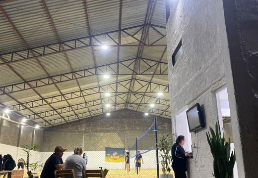 Área de convivência com mesas de madeira e TV ao lado das quadras.