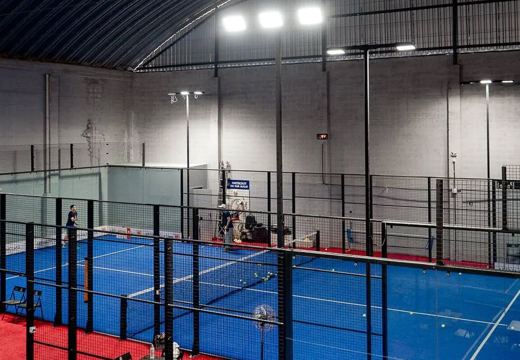 Quadra de padel coberta com piso azul, paredes de vidro, proteção metálica e iluminação.