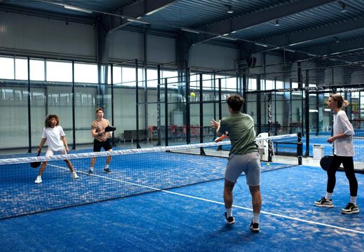 Jogadores em quadra de padel coberta, com piso azul, paredes de vidro e iluminação.