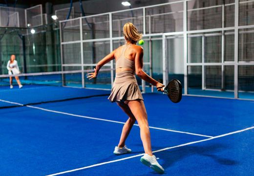 Partida de padel em quadra de piso azul, com fechamento de vidro e proteção metálica.