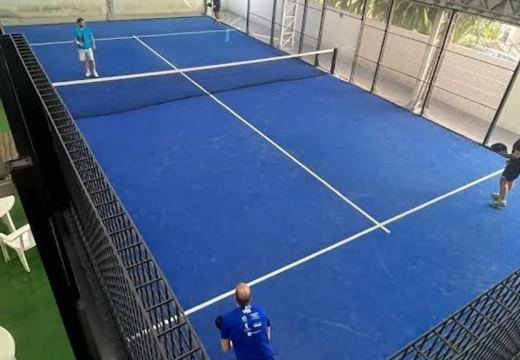 Vista da quadra de padel coberta, com piso azul, rede, iluminação e proteção perimetral.