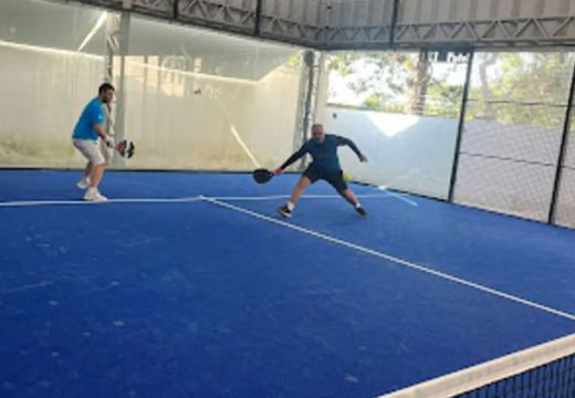 Jogadores em uma quadra de padel de piso azul, cercada por vidro e estrutura metálica.