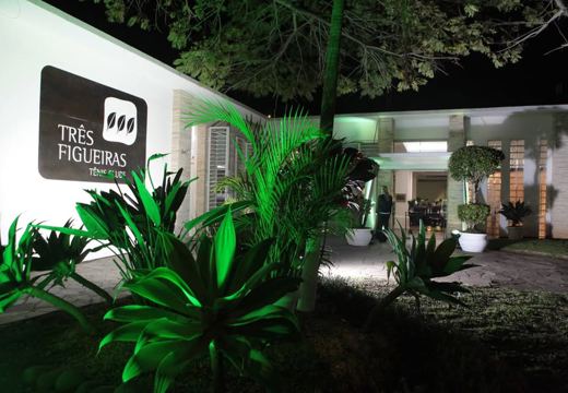 Entrada do Três Figueiras Tênis Clube à noite, com a marca na fachada e jardim iluminado.