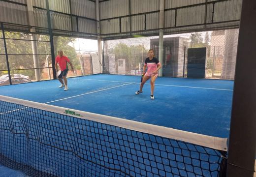 Partida em quadra de padel coberta, com piso azul, paredes de vidro e proteção metálica.