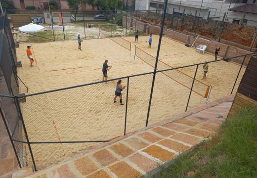 Vista das quadras de beach tennis de areia em sua configuração anterior, ao ar livre, com redes e proteção perimetral.